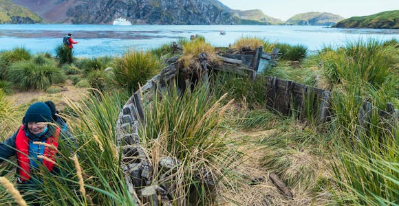 A hiker walks past an abandoned wooden boat in the grass at Godthul bay in South Georgia