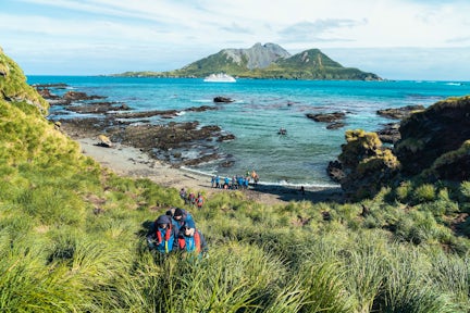 A group of tourists climb a grassy hill up from a beach at Cooper Bay in South Georgia. A zodiac drives to a cruise ship is anchored in the bay.