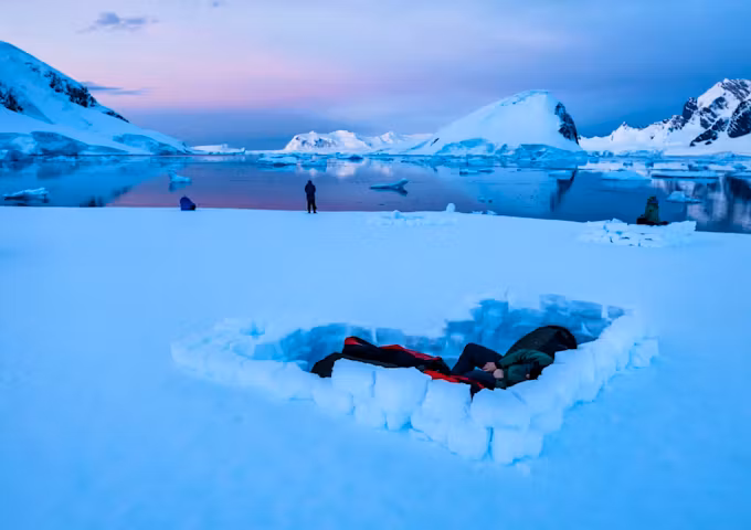 Campers camp in a snowhole in Antarctica while a pink-tinged sun sets in the background