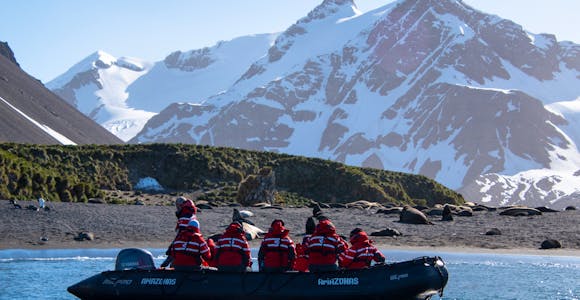 A zodiac boat full of tourists in red jackets cruises in Right Whale Bay in South Georgia, with snowy mountains in the background
