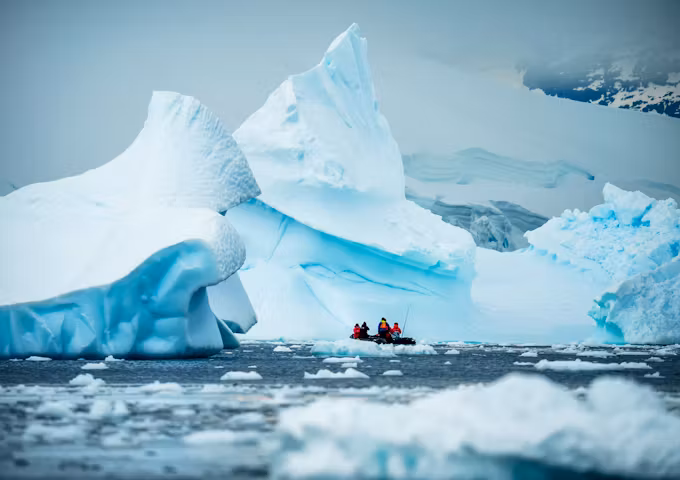 A zodiac rib boat cruises through an icy Antarctic sea with a huge ice formation in the background