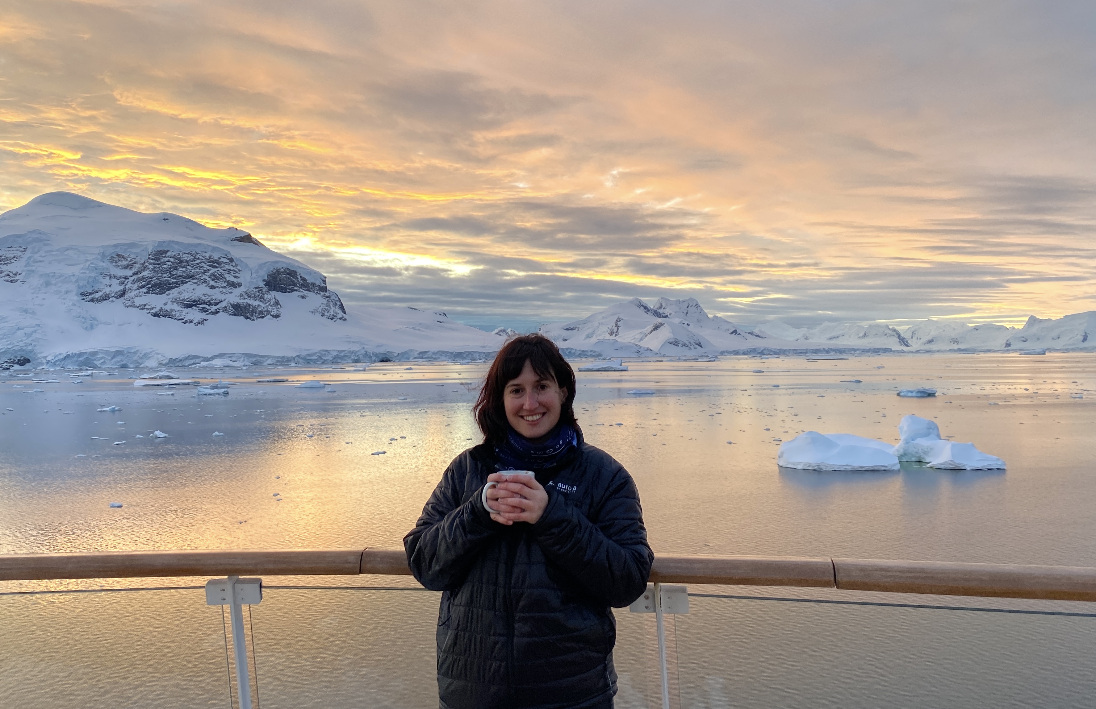 Carys from Swoop stands on the deck of a ship while holding a cup. She is smiling while the sun sets in the Antarctic background