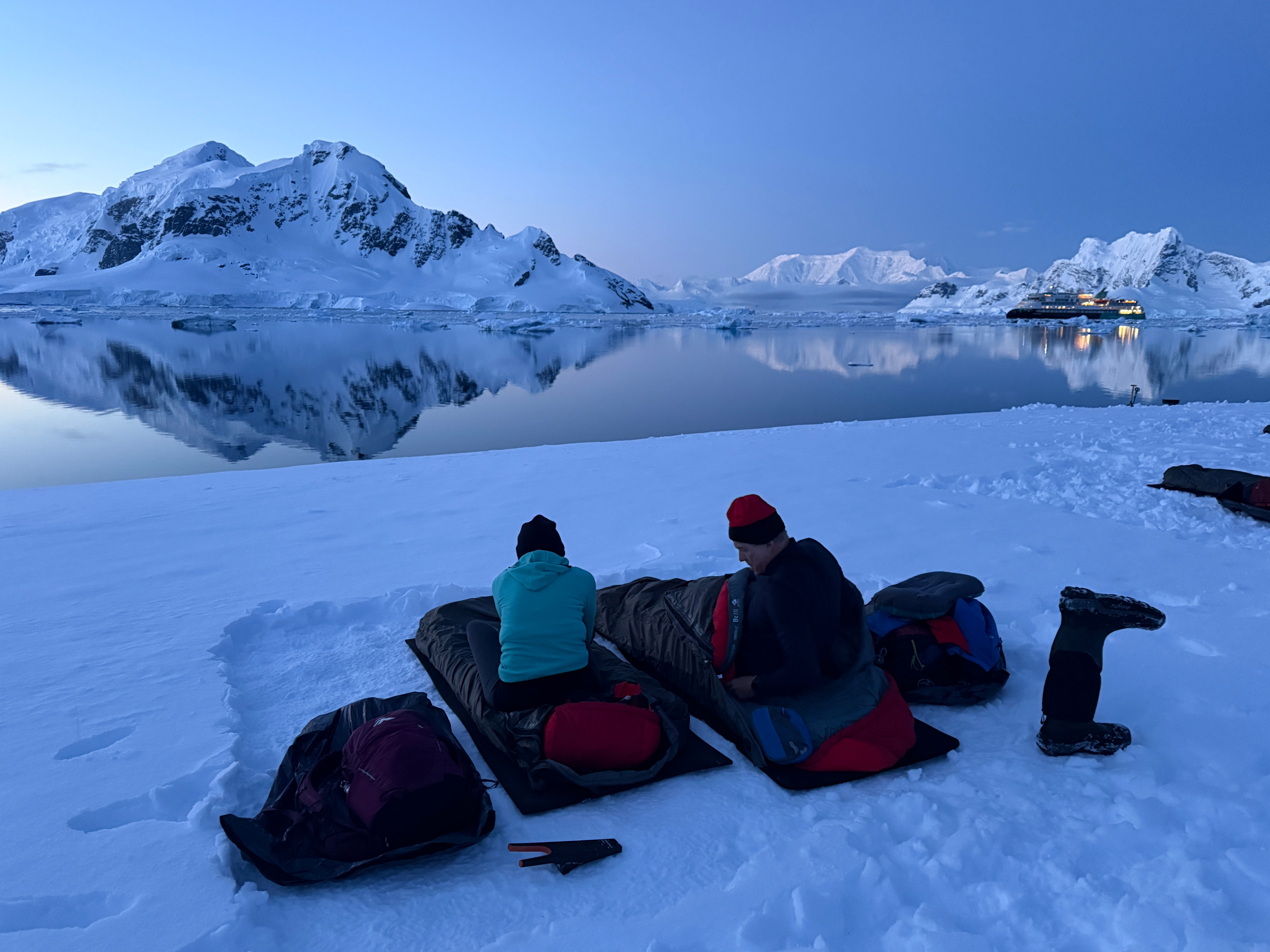 Two people sit on sleeping bags in a dusky Antarctic bay ahead of a night camping