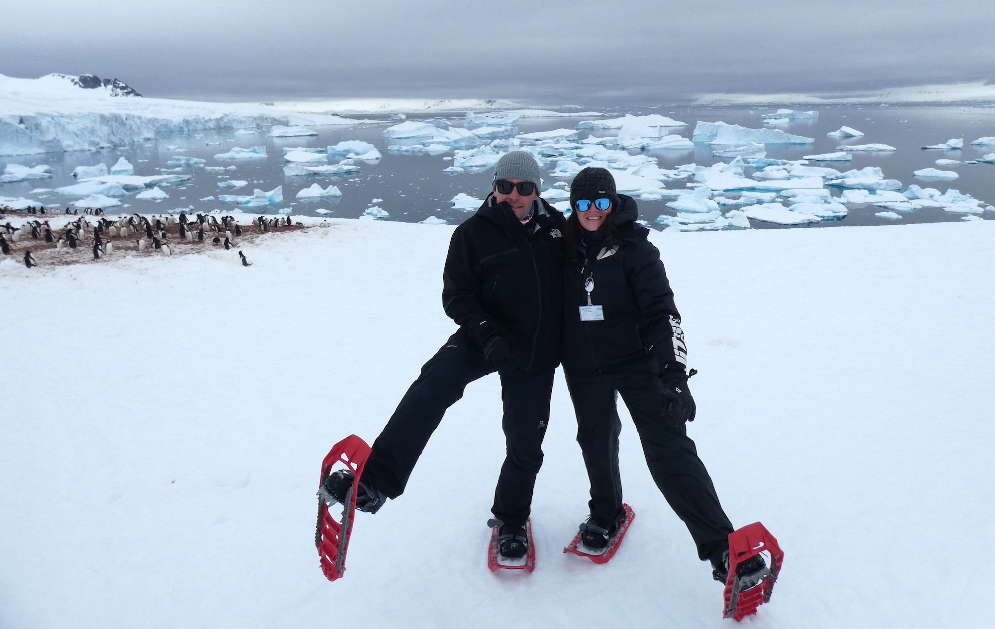 A man and woman show off their snowshoes amid a snowy Antarctic backdrop