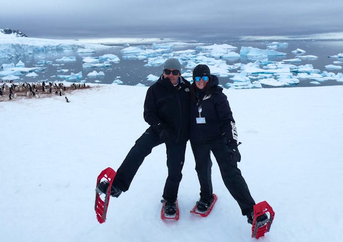 A man and woman show off their snowshoes amid a snowy Antarctic backdrop