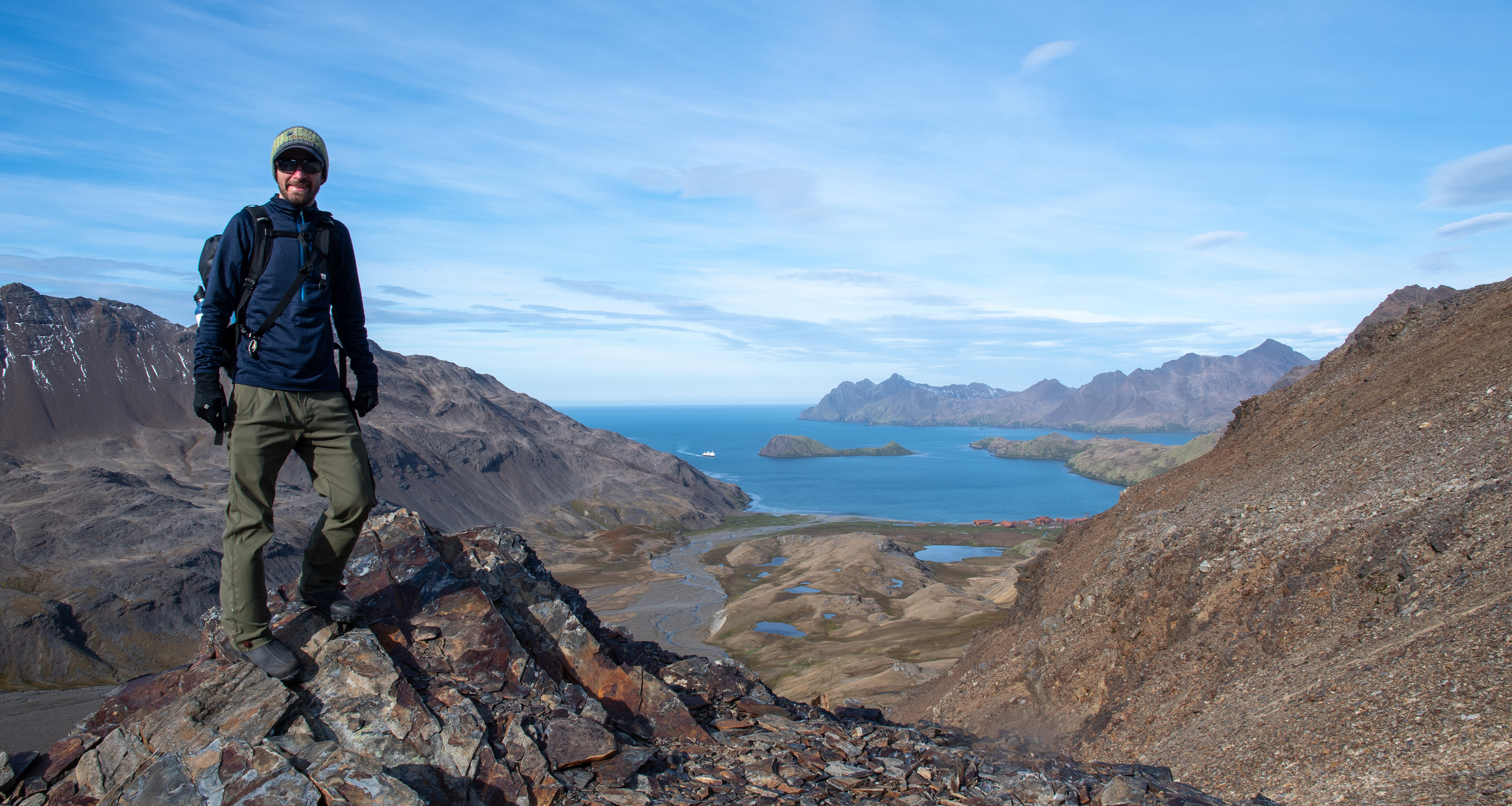 A man stands on a mountain ridge on South Georgia Island. Behind him is a distant blue bay with Stromness whaling station, at the end of the Shackleton hike