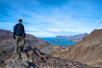 A man stands on a mountain ridge on South Georgia Island, Behind him a ridge descends to a distant blue bay, with Stromness whaling station just visible at the end of the Shackleton hike