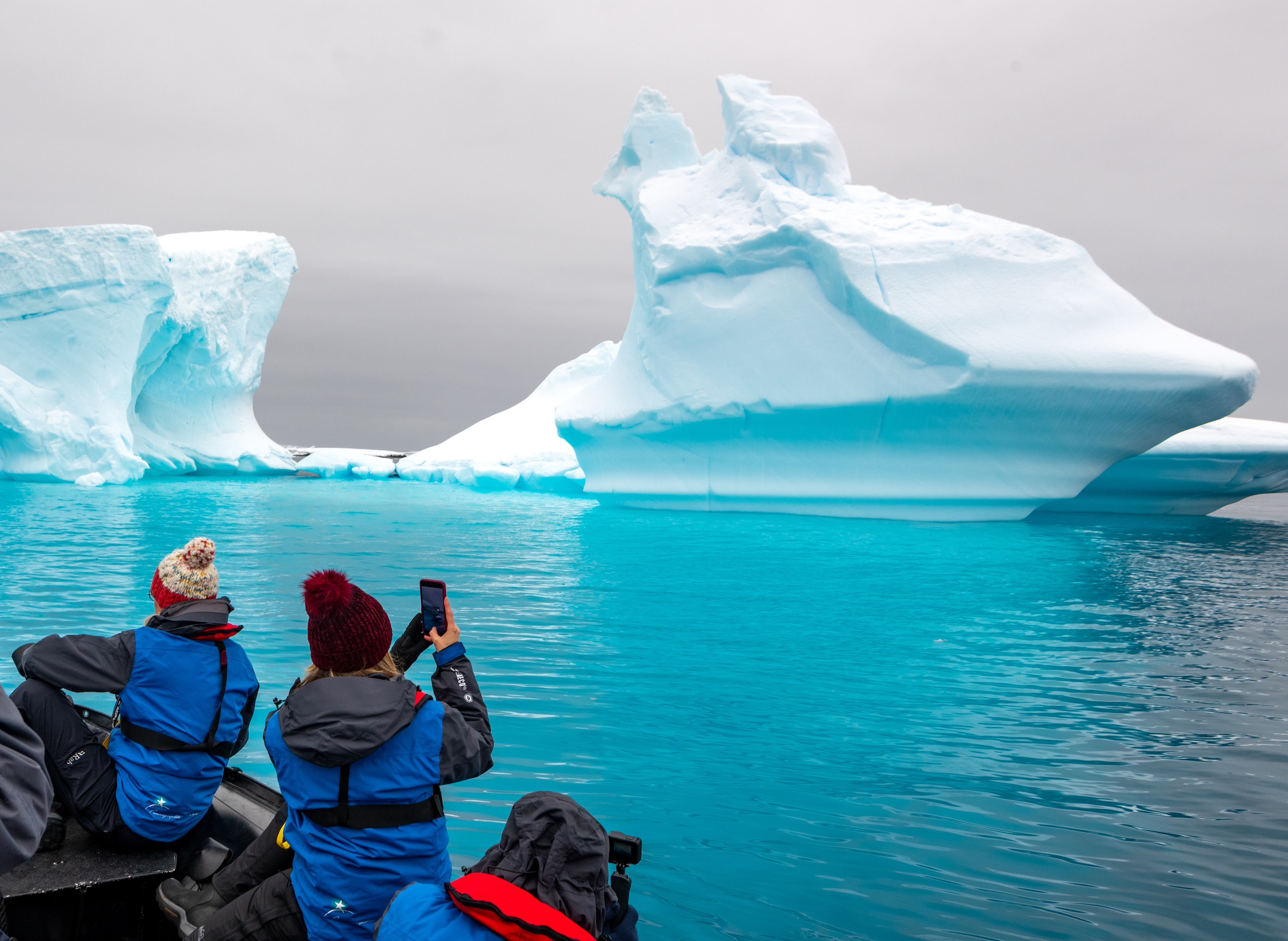 Three people perch on the side of a boat in Antarctica in front of an ice formation and with bright turquoise water just below