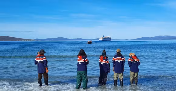 A team of guides waits for a zodiac of passengers to land on a beach at Saunders Island in the Falkland Islands. Their cruise ship is in the background